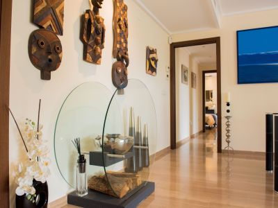 Luminoso pasillo con suelo de gres porcelánico brillante efecto madera, decorado con máscaras tribales de madera y un moderno expositor de cristal con leña y orquídeas. Un cuadro abstracto azul completa la decoración.