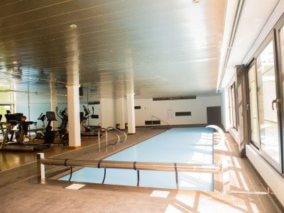 Piscina cubierta con cubierta protectora, rodeada de tarima de madera. Grandes ventanales inundan el espacio de luz natural, y un gimnasio se refleja en el espejo del fondo.