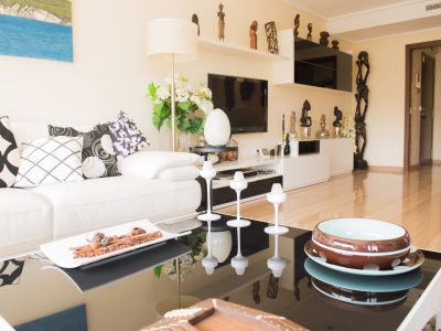 Salón con sofá blanco, cojines gráficos y mesa de centro negra brillante con detalles decorativos. Mueble de TV con estantes y esculturas africanas.