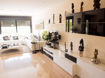Salón luminoso con sofá seccional blanco, suelo de baldosas pulidas y mueble de TV moderno. La luz natural entra por la ventana con persianas enrollables.