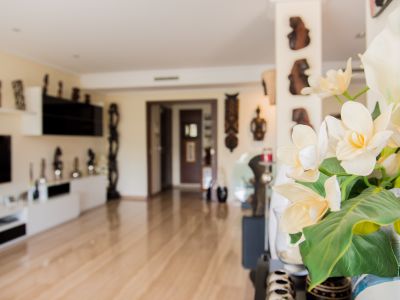 Vaso en primer plano con elegantes flores artificiales blancas y amarillas. Interior luminoso con suelo de madera clara, que conduce a una moderna sala de estar con arte mural decorativo.
