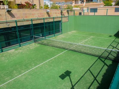 Vista de la pista de pádel exterior de la comunidad, con césped artificial verde vibrante, red y robustas paredes de malla verde, y edificios residenciales al fondo soleado.
