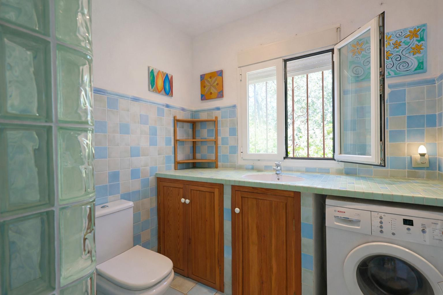 Baño funcional con azulejos en tonos azules, encimera alicatada con lavabo integrado, armarios de madera y zona de lavandería con luz natural.