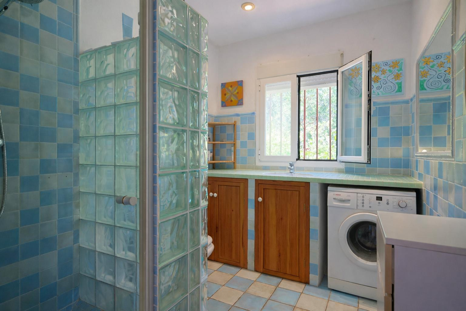 Cuarto de baño funcional con zona de lavandería, azulejos en tonos azules, ducha con pavés de vidrio, encimera alicatada y mobiliario de madera natural.
