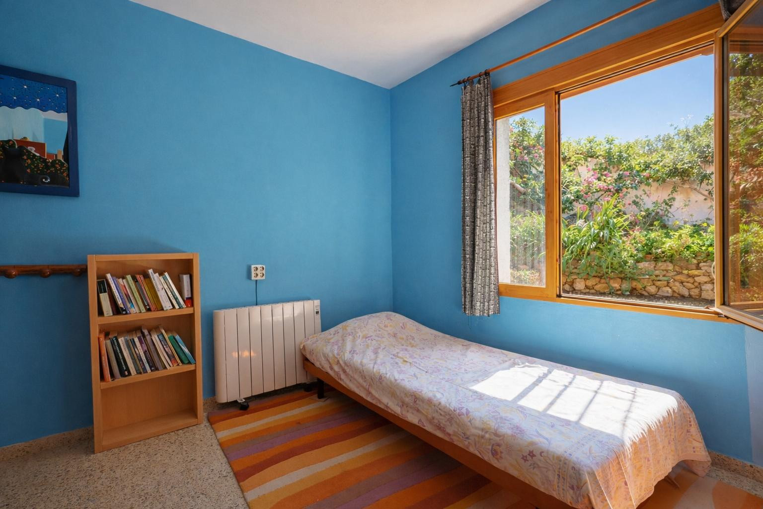 Dormitorio con paredes en azul intenso, cama individual y estantería de madera. La ventana de madera ofrece vistas directas al jardín y muros de piedra natural.