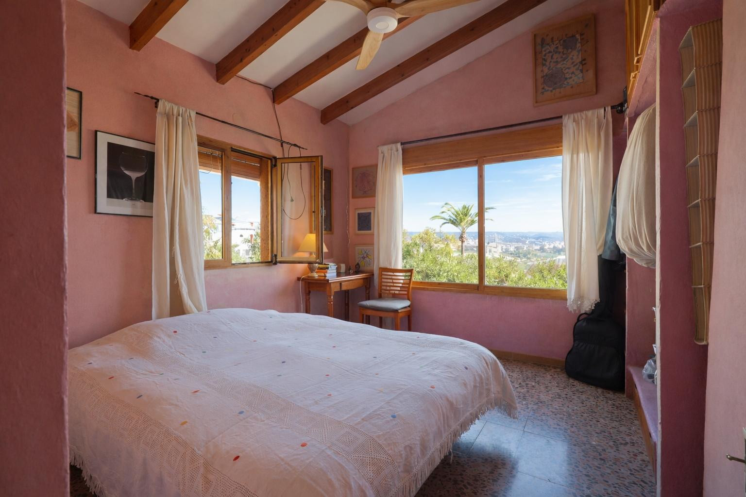 Dormitorio con techos de vigas de madera, paredes en tonos cálidos y amplios ventanales con vistas despejadas al entorno natural de Jávea.