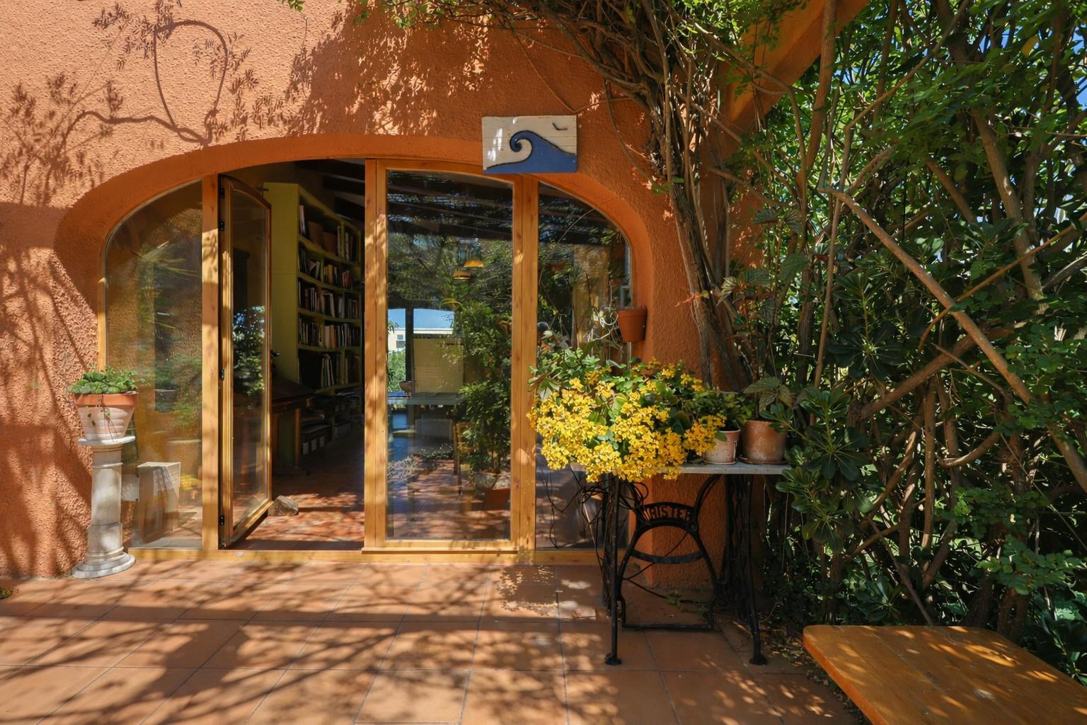 Entrada exterior con carpintería de madera arqueada y puertas acristaladas. Destacan el suelo de terracota y la vegetación integrada que refresca el acceso a la vivienda.