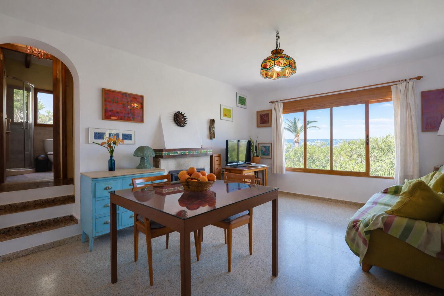Salón comedor con chimenea decorativa, suelos de terrazo y un gran ventanal de madera con vistas al mar. Un arco lateral comunica con el baño de la vivienda.