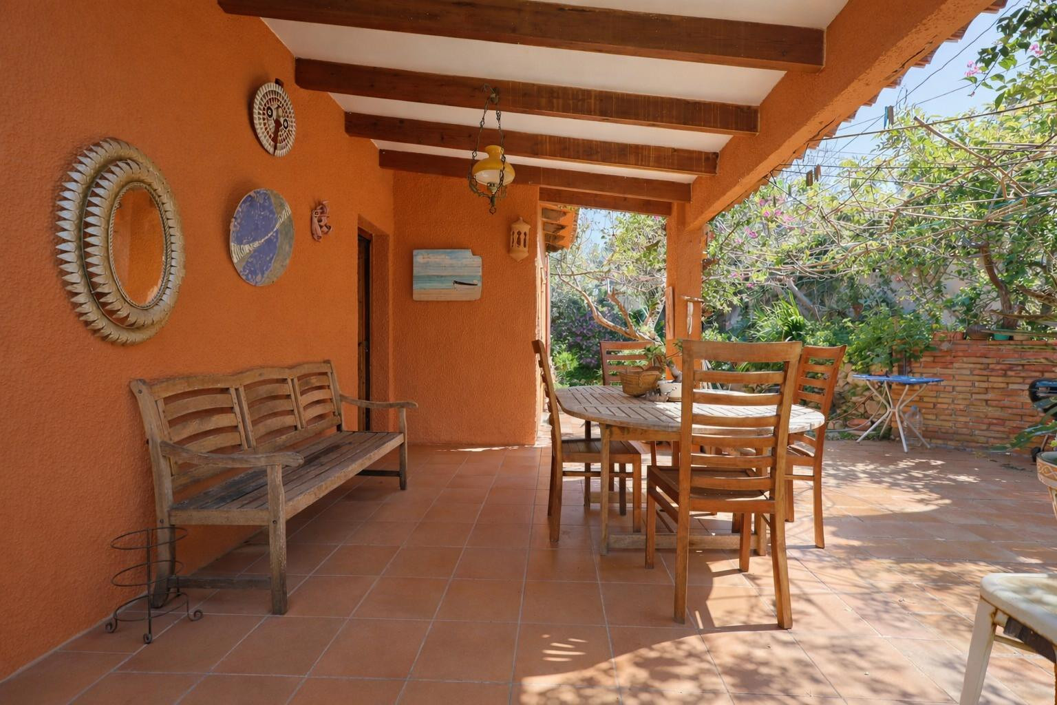 Terraza cubierta con vigas de madera a la vista, suelos de terracota y mobiliario de comedor exterior rodeado de paredes en tonos tierra.