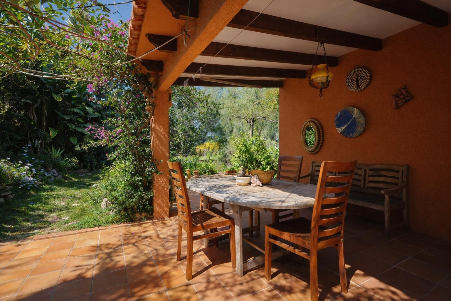 Terraza cubierta con vigas de madera vista, suelo de terracota y comedor exterior rodeado de un jardín mediterráneo maduro con buganvillas.