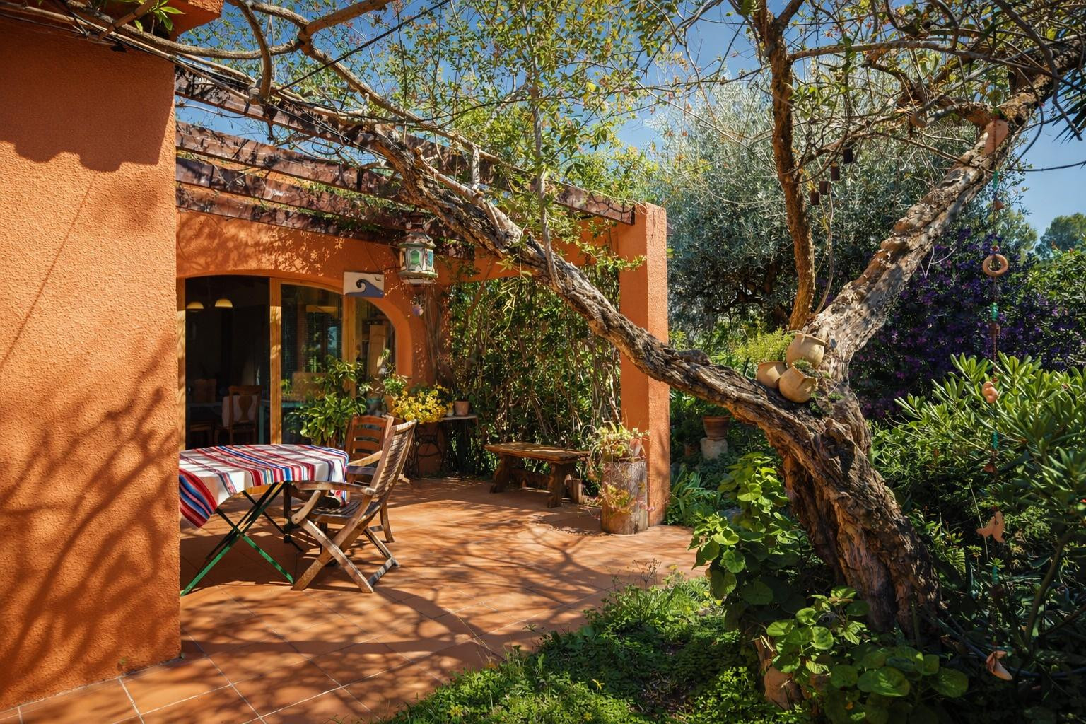 Terraza exterior con suelo de barro cocido, pérgola de madera y vegetación madura que proporciona sombra natural sobre el área de comedor.