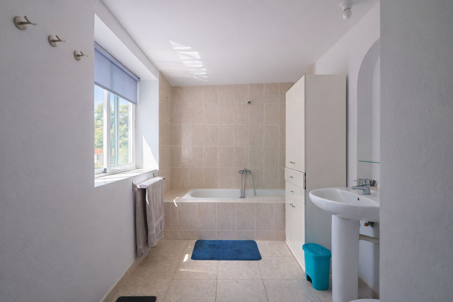 Baño funcional con bañera integrada, revestimiento cerámico en tonos neutros, lavabo de pedestal y ventana con vistas al exterior.