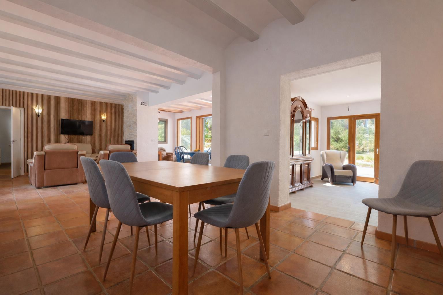 Comedor con mesa de madera y sillas tapizadas, suelos de terracota y techos con vigas blancas que conectan con el salón y las estancias luminosas.