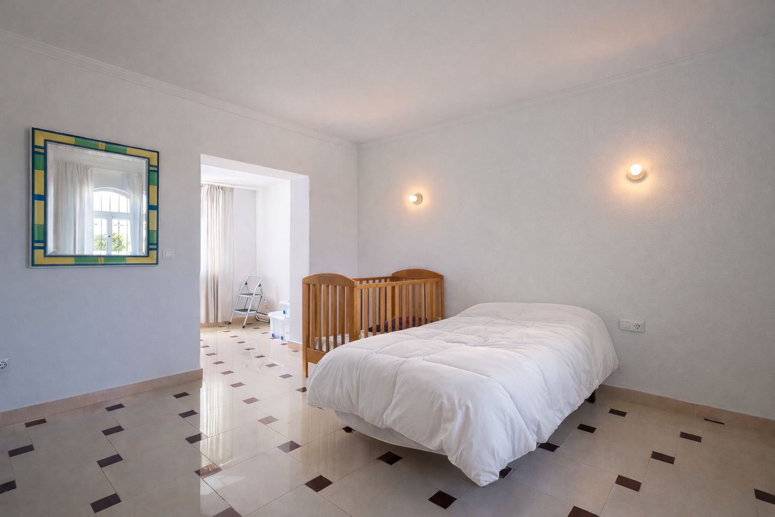 Dormitorio luminoso con suelos de gres decorados, cama individual y cuna de madera. Incluye apliques de pared y acceso a una estancia contigua con luz natural.