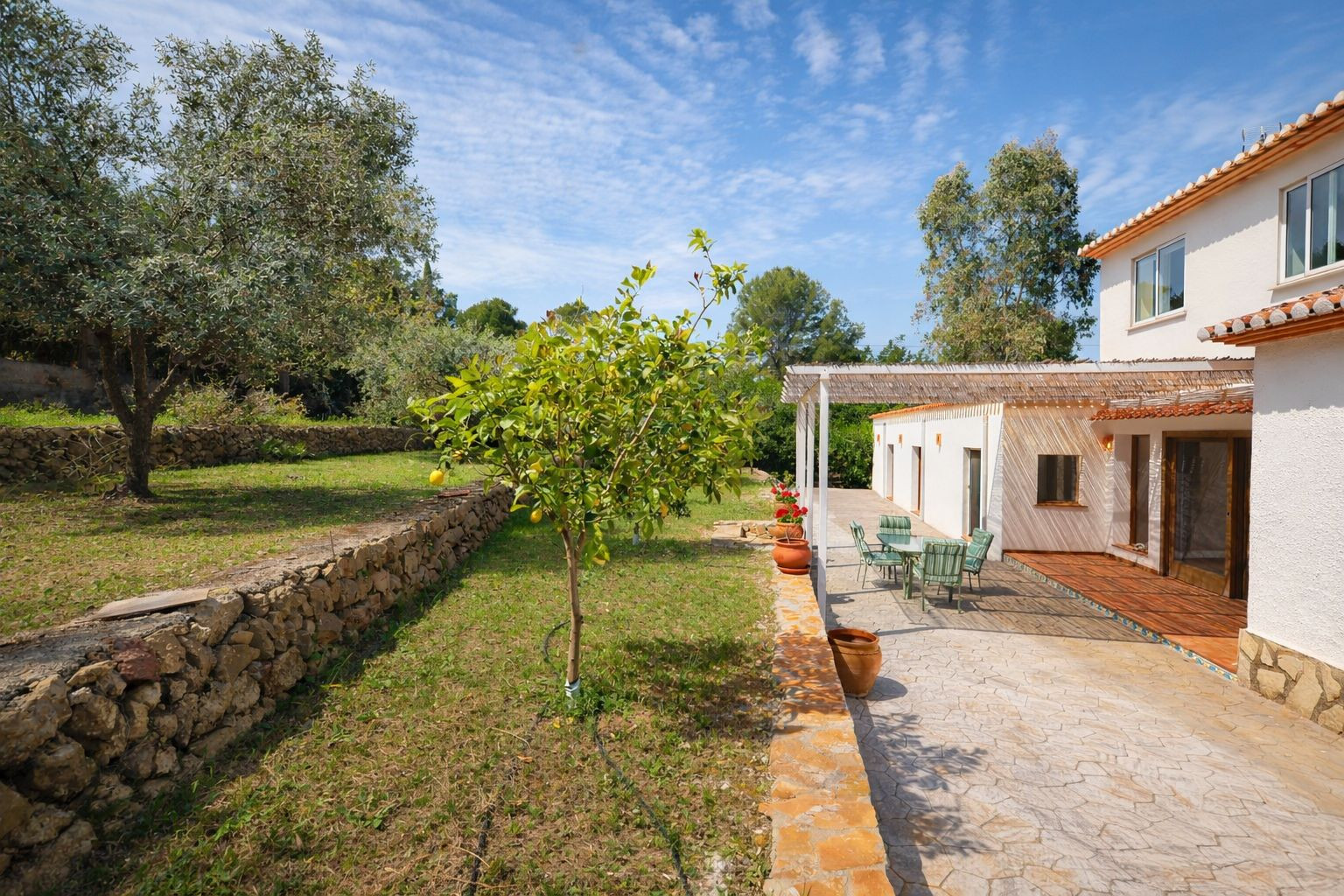 Exterior con terraza pavimentada, pérgola de sombra y jardín aterrazado con muros de piedra natural y árboles frutales bajo un cielo despejado.