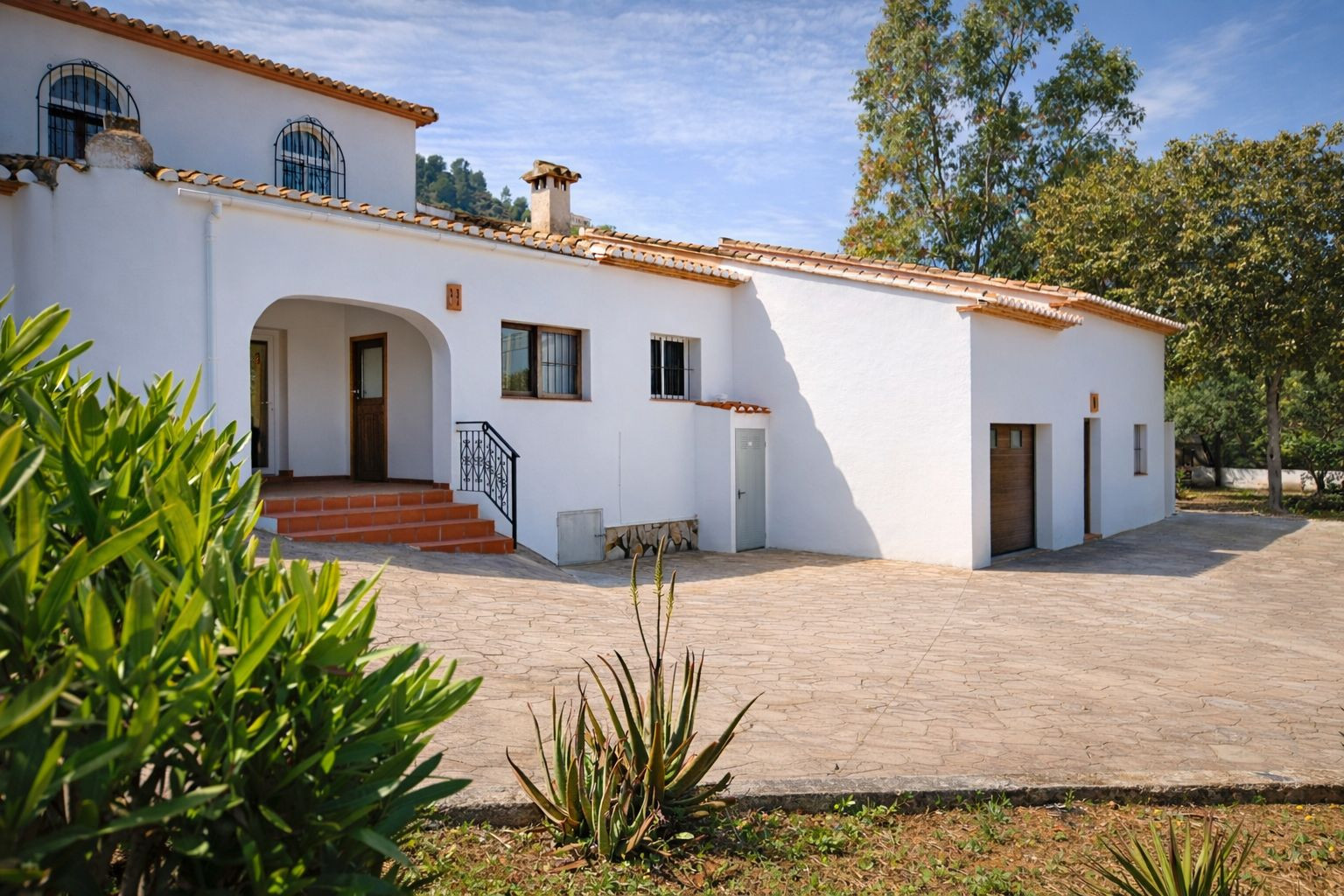 Fachada blanca de estilo mediterráneo con tejado de teja cerámica, porche de entrada arqueado y amplio patio pavimentado con acceso a garaje integrado.