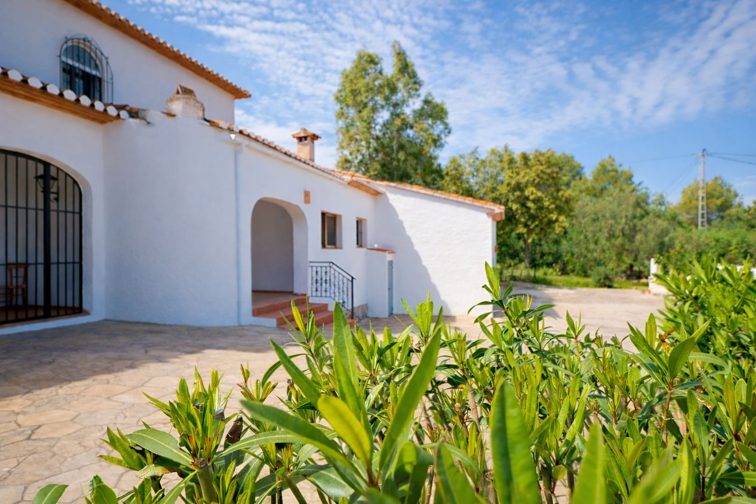 Fachada exterior de la villa con paredes blancas, tejado de teja cerámica y patio de piedra natural. Se observa un acceso arqueado y vegetación mediterránea.