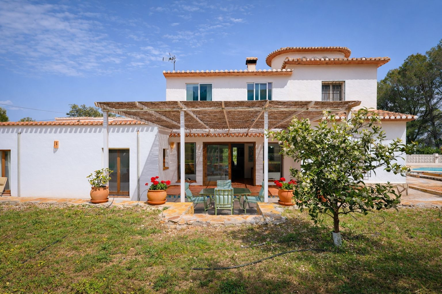 Fachada posterior de la villa con porche cubierto por cañizo, zona de comedor exterior y jardín con árboles frutales bajo un cielo despejado.