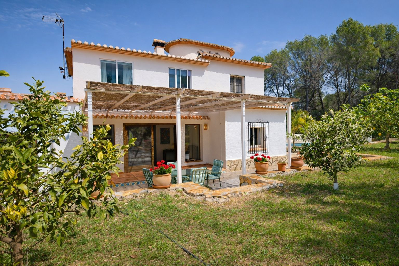 Fachada posterior de la villa con porche de cañizo, zona de estar exterior y jardín con frutales. Destaca la arquitectura mediterránea con acabados en piedra y teja.