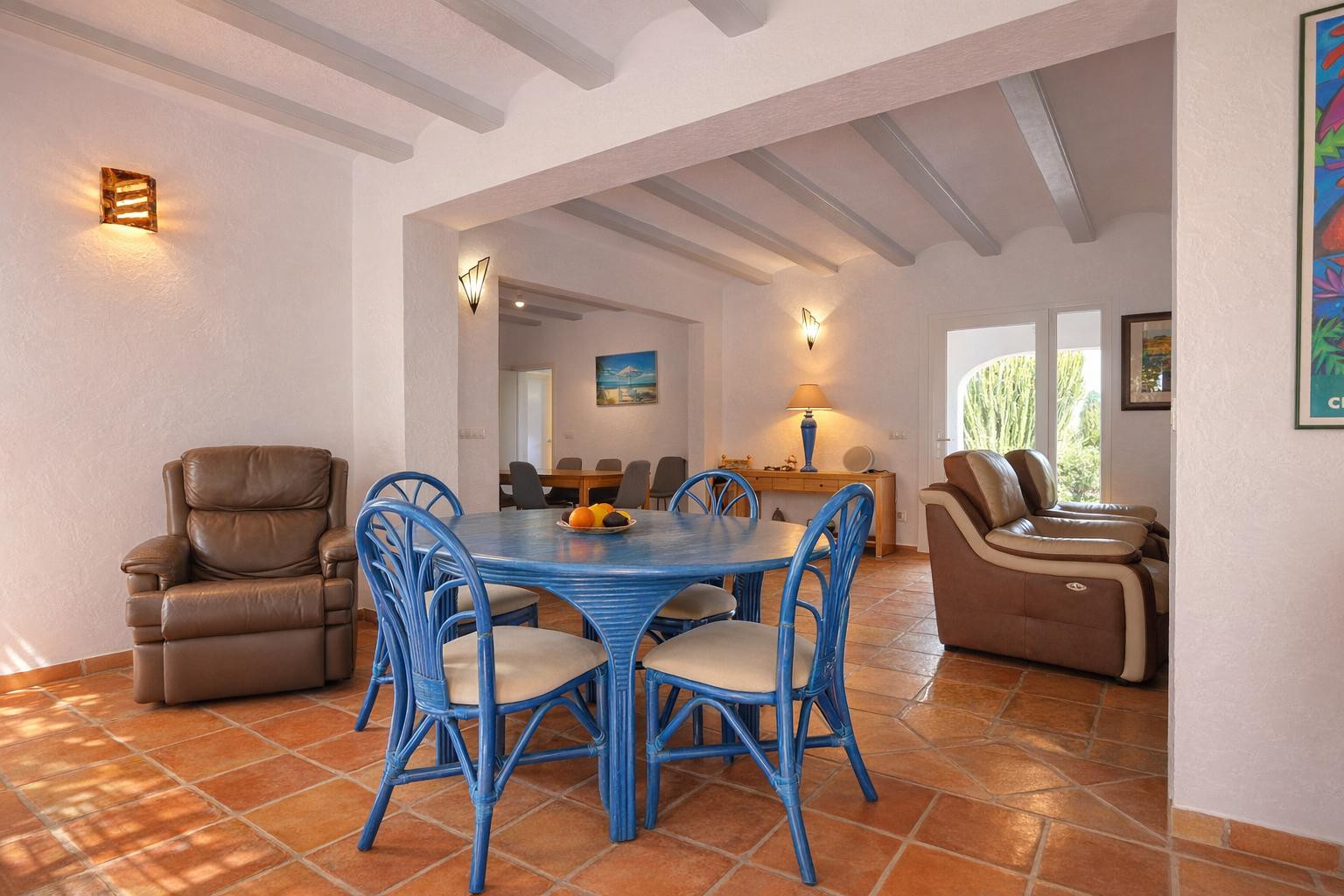 Salón-comedor con suelos de terracota, vigas de madera a la vista en blanco y mobiliario de mimbre azul. El espacio abierto conecta con una segunda zona de estar luminosa.