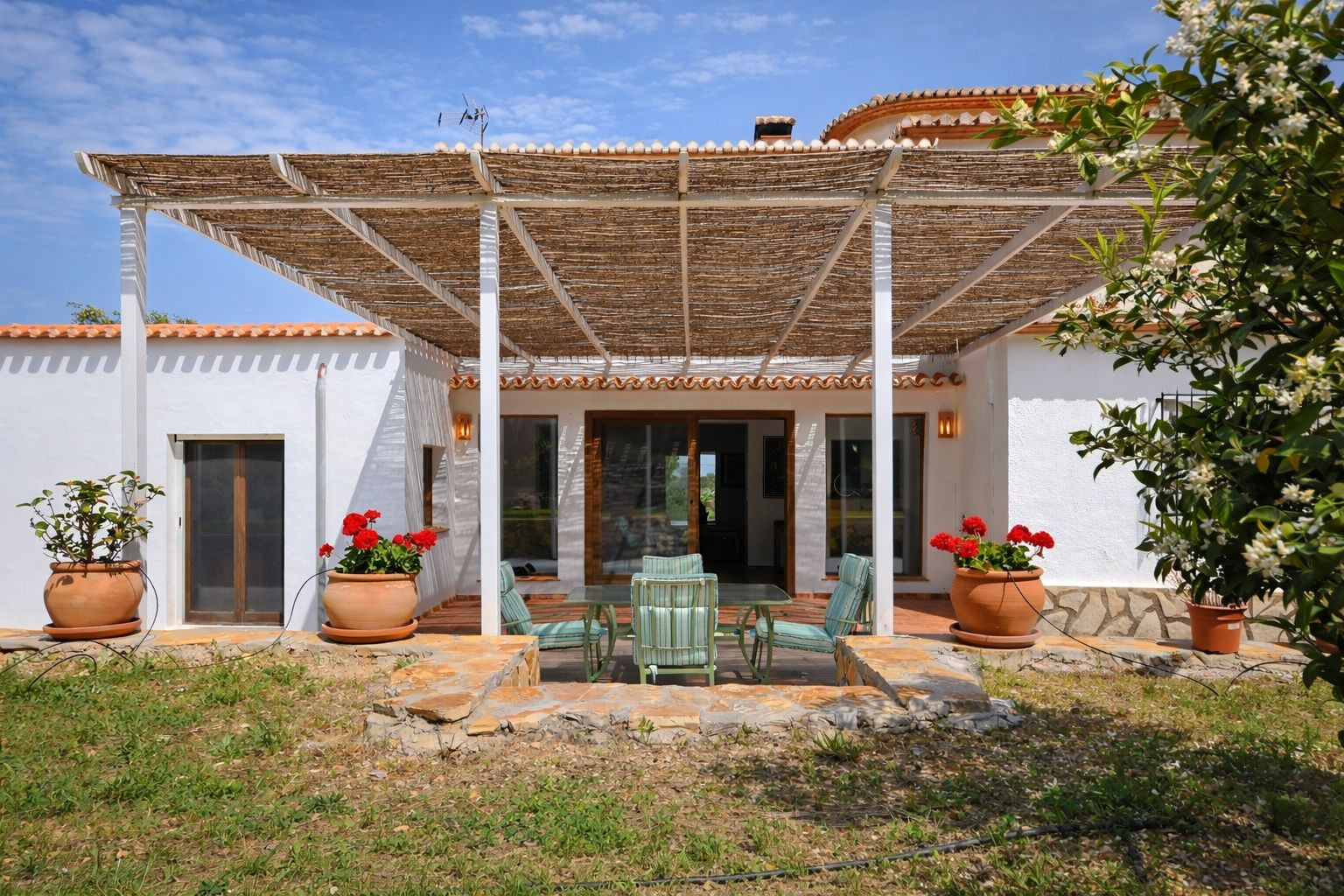 Terraza exterior con pérgola de cañizo, mobiliario de comedor y macetas de terracota con geranios rojos sobre suelo de piedra natural.