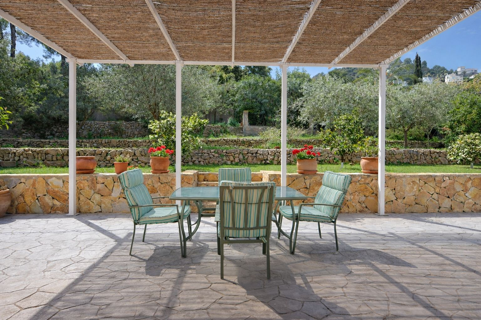 Terraza exterior con pérgola de cañizo, mobiliario de comedor y vistas a un jardín abancalado con muros de piedra natural y árboles frutales.