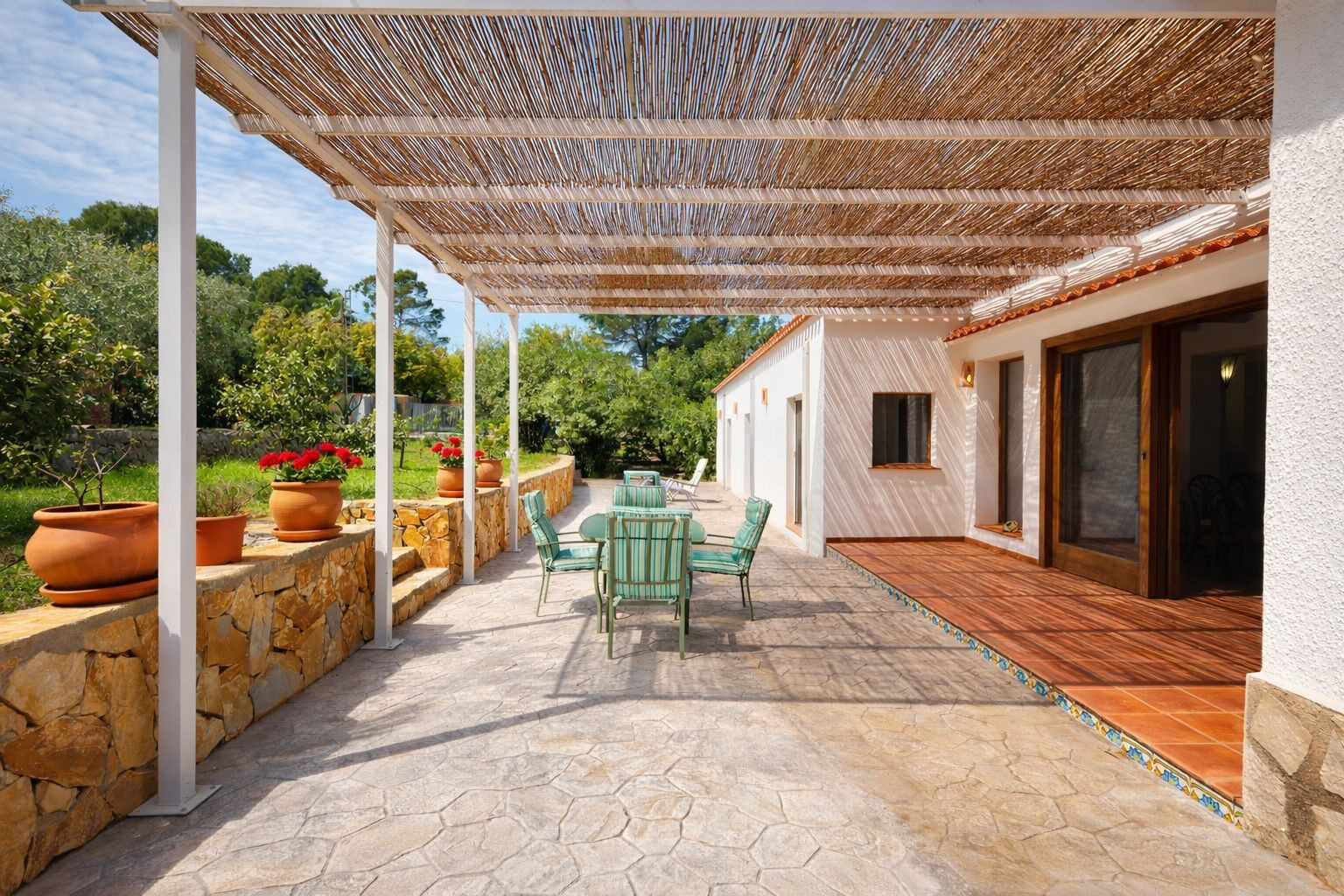 Terraza exterior con pérgola de cañizo, suelo de piedra y mobiliario de jardín. Incluye un murete de piedra natural con macetas y vistas al jardín frutal.