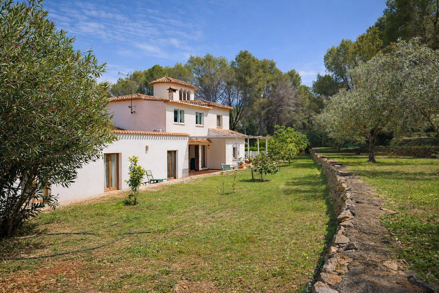 Vista exterior de la villa con fachada blanca y tejado de teja árabe, rodeada de un jardín con árboles frutales y un muro perimetral de piedra natural.