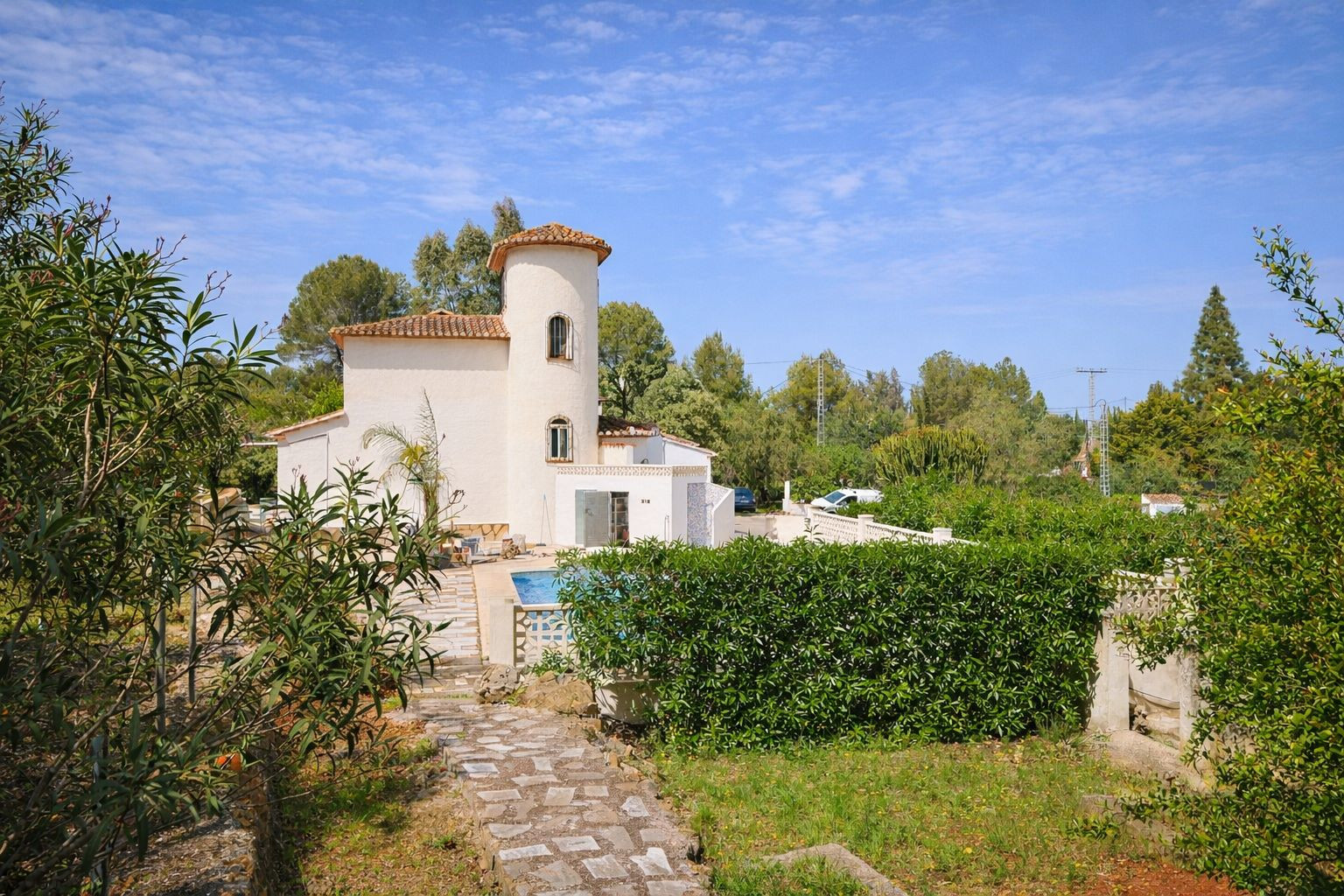 Vista exterior de la villa con torre circular, tejado de teja árabe y jardín mediterráneo con senderos de piedra y piscina privada.