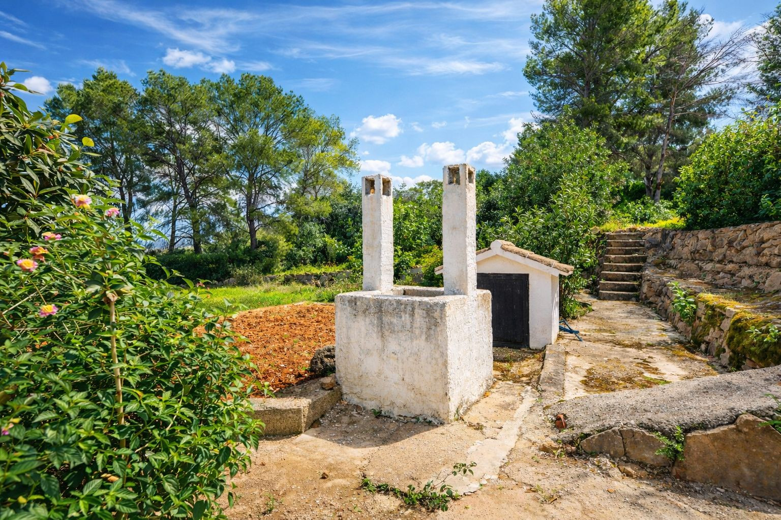 Zona exterior rústica con barbacoa de mampostería blanca, chimeneas dobles y escaleras de piedra que acceden a la zona superior ajardinada rodeada de pinos.