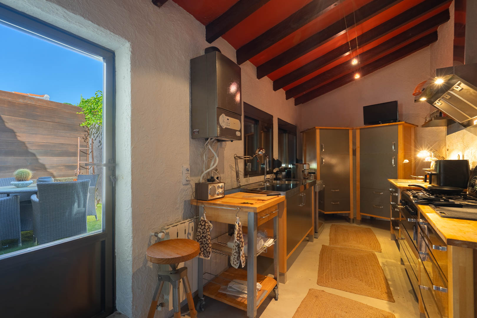 Cocina rústica con vigas de madera oscura, encimeras de madera y acceso directo a un patio exterior privado a través de una amplia puerta acristalada.