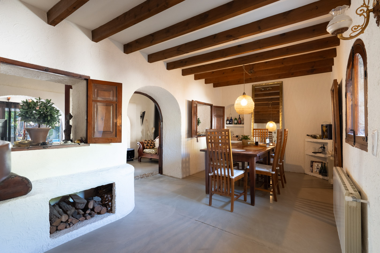 Comedor rústico con vigas de madera vista, mesa de madera maciza y un arco que conecta con el salón. Destaca el leñero integrado y las ventanas interiores con contraventanas de madera.