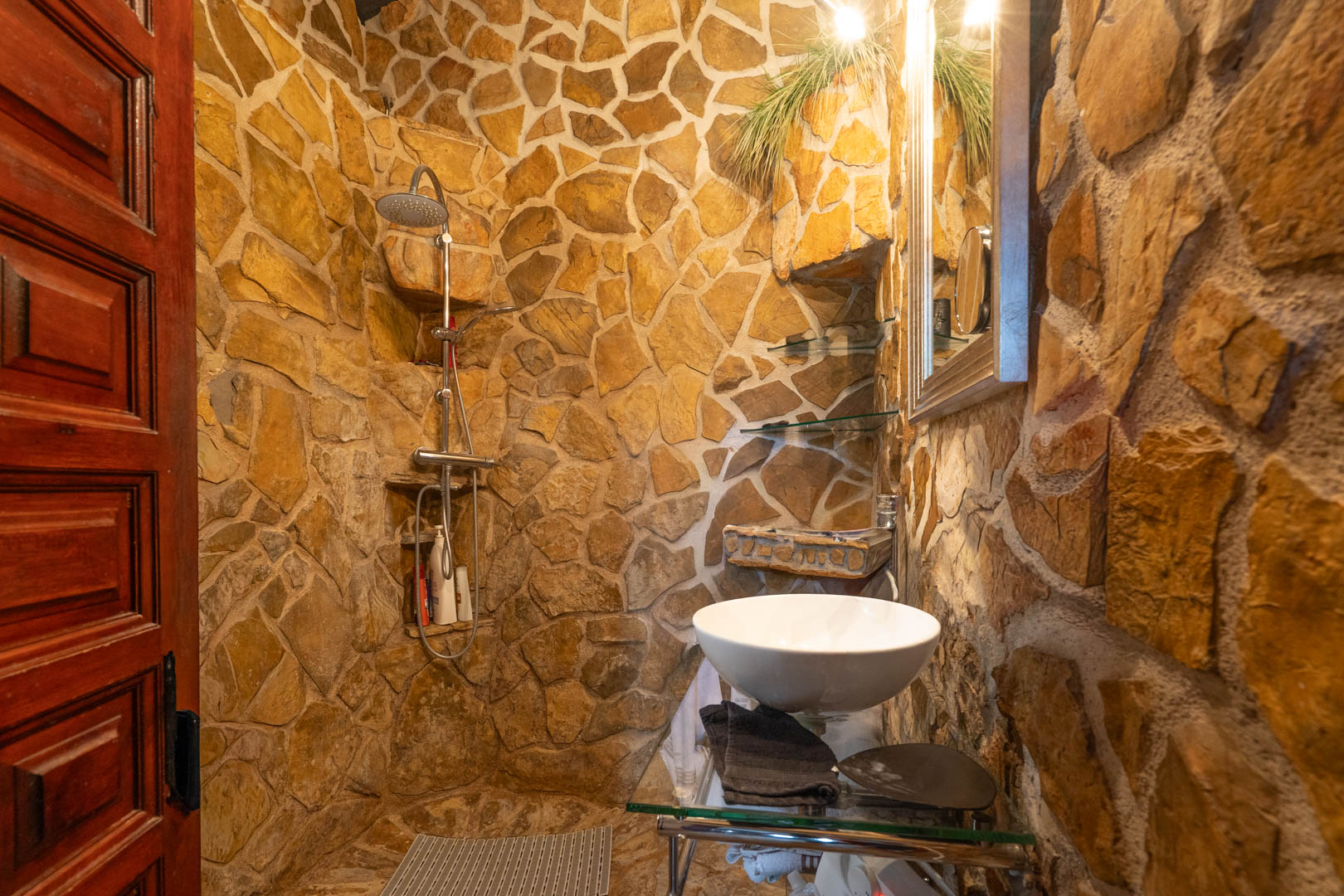 Cuarto de baño rústico con paredes de piedra natural, zona de ducha integrada con estantes de obra y lavabo sobre encimera de cristal.