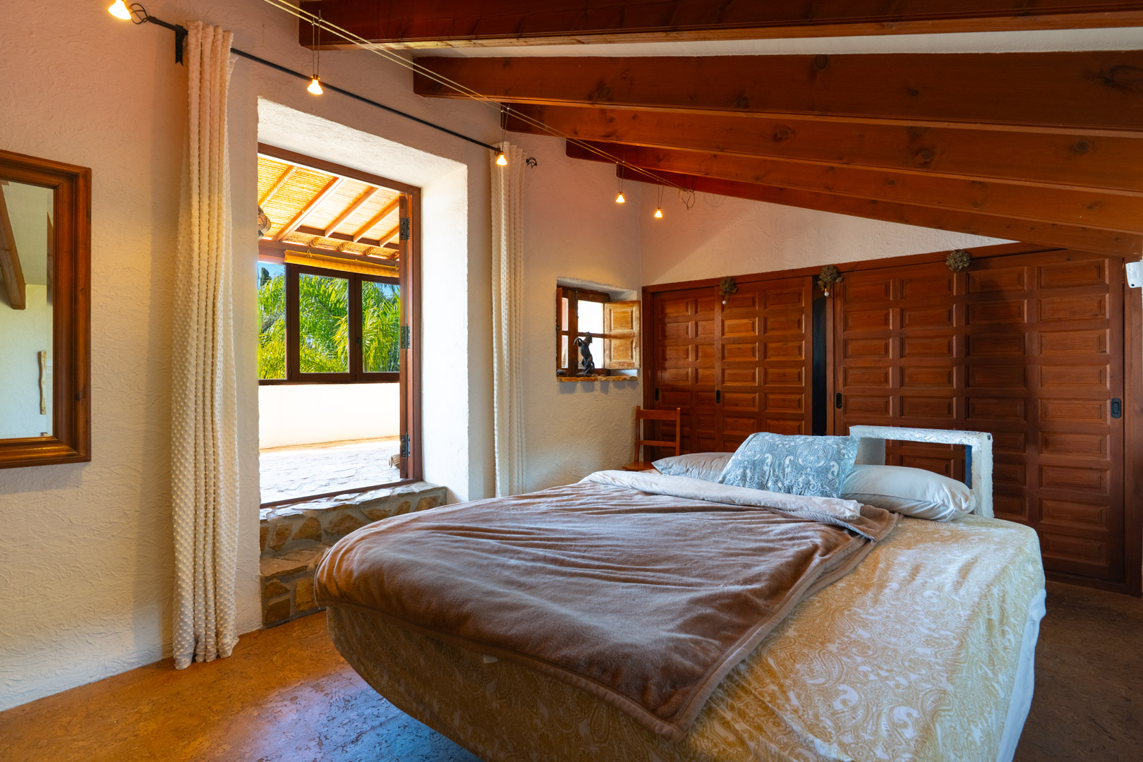 Dormitorio con techos de vigas de madera, armarios empotrados de estilo rústico y acceso a una terraza con vistas despejadas al entorno natural.