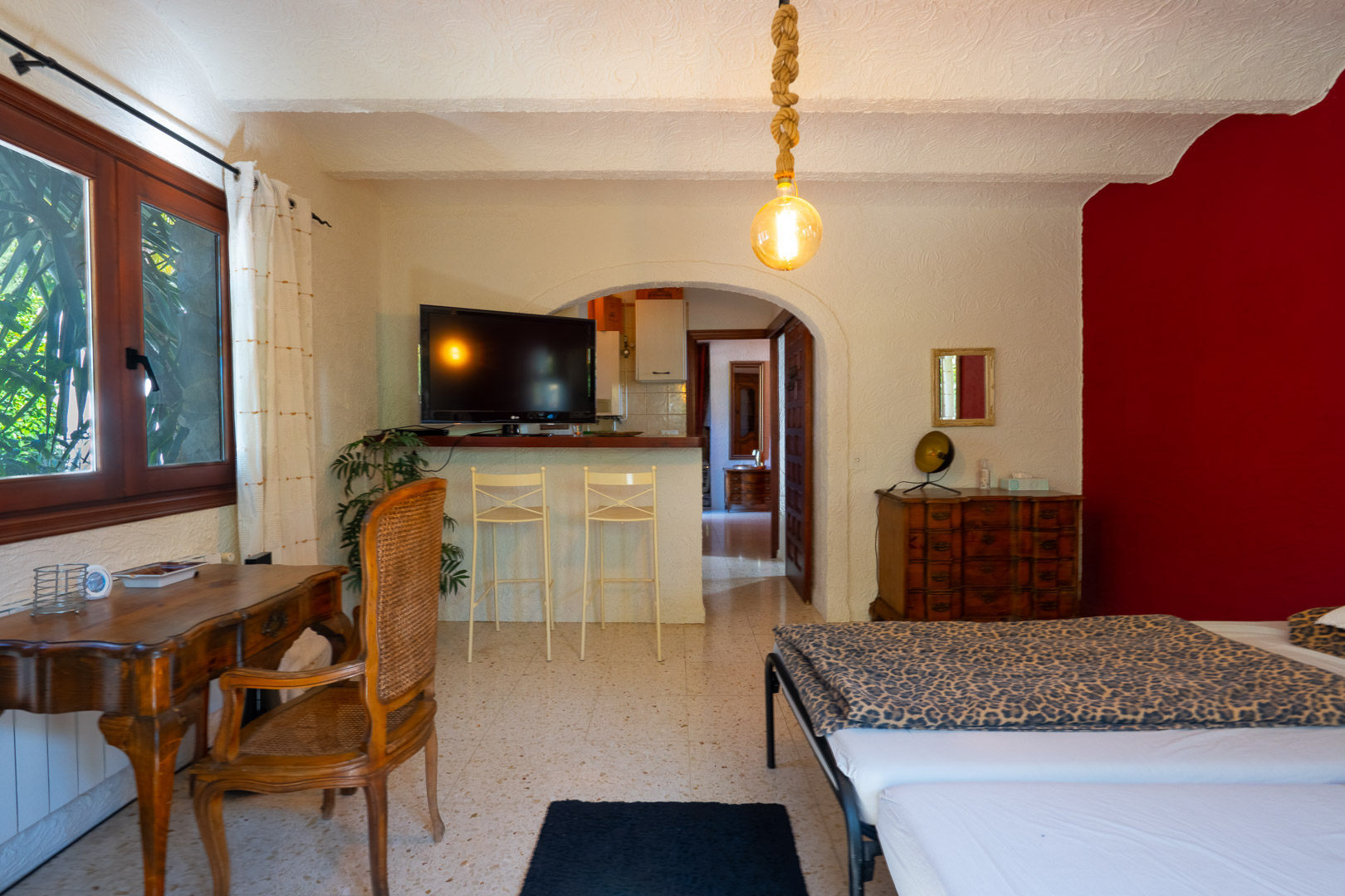 Dormitorio de invitados con zona de estar, escritorio de madera clásica, pared de acento roja y barra con taburetes que conecta con la cocina a través de un arco.
