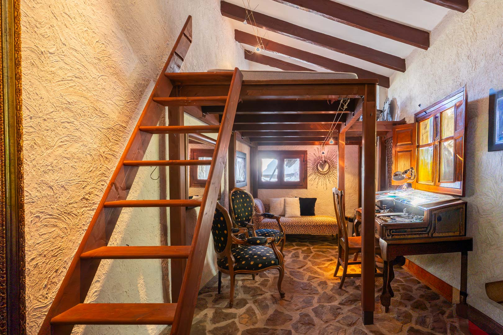 Habitación rústica con altillo de madera, vigas vistas y suelo de piedra natural. Incluye una zona de despacho con escritorio clásico y mobiliario de estilo vintage.