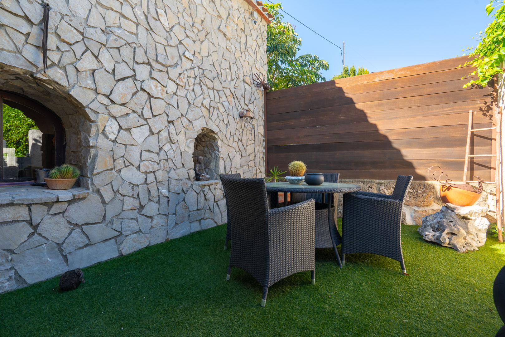 Patio exterior con muro de mampostería de piedra natural, conjunto de comedor de ratán oscuro sobre césped artificial y hornacinas decorativas.