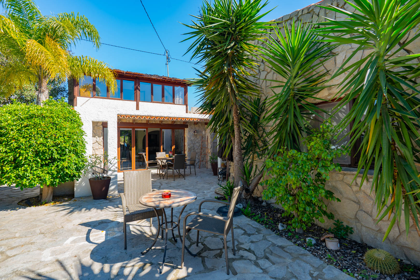 Patio exterior con suelo de piedra natural, vegetación mediterránea y fachada que combina muros de piedra con una naya acristalada en la planta superior.