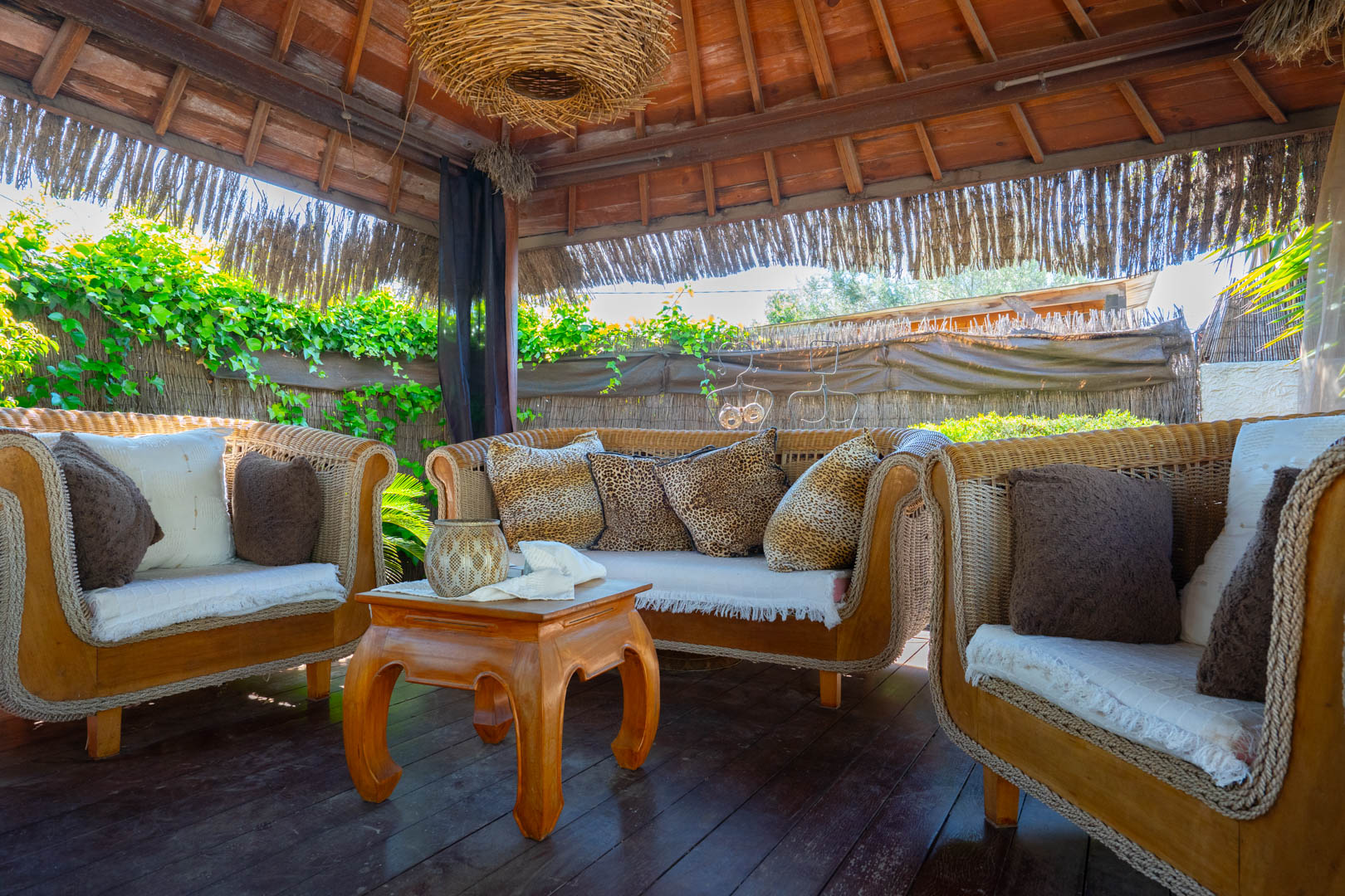 Pérgola balinesa con techo de madera y paja, equipada con sofás de mimbre, cojines decorativos y una mesa de centro sobre suelo de tarima exterior.