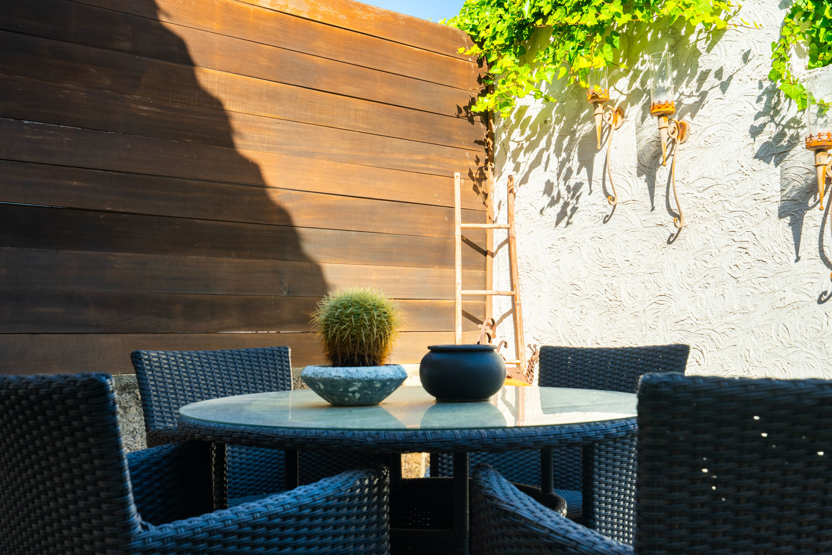 Rincón exterior con mesa redonda de cristal y sillas de mimbre oscuro, decorado con cactus y apliques de forja sobre una pared de textura blanca.