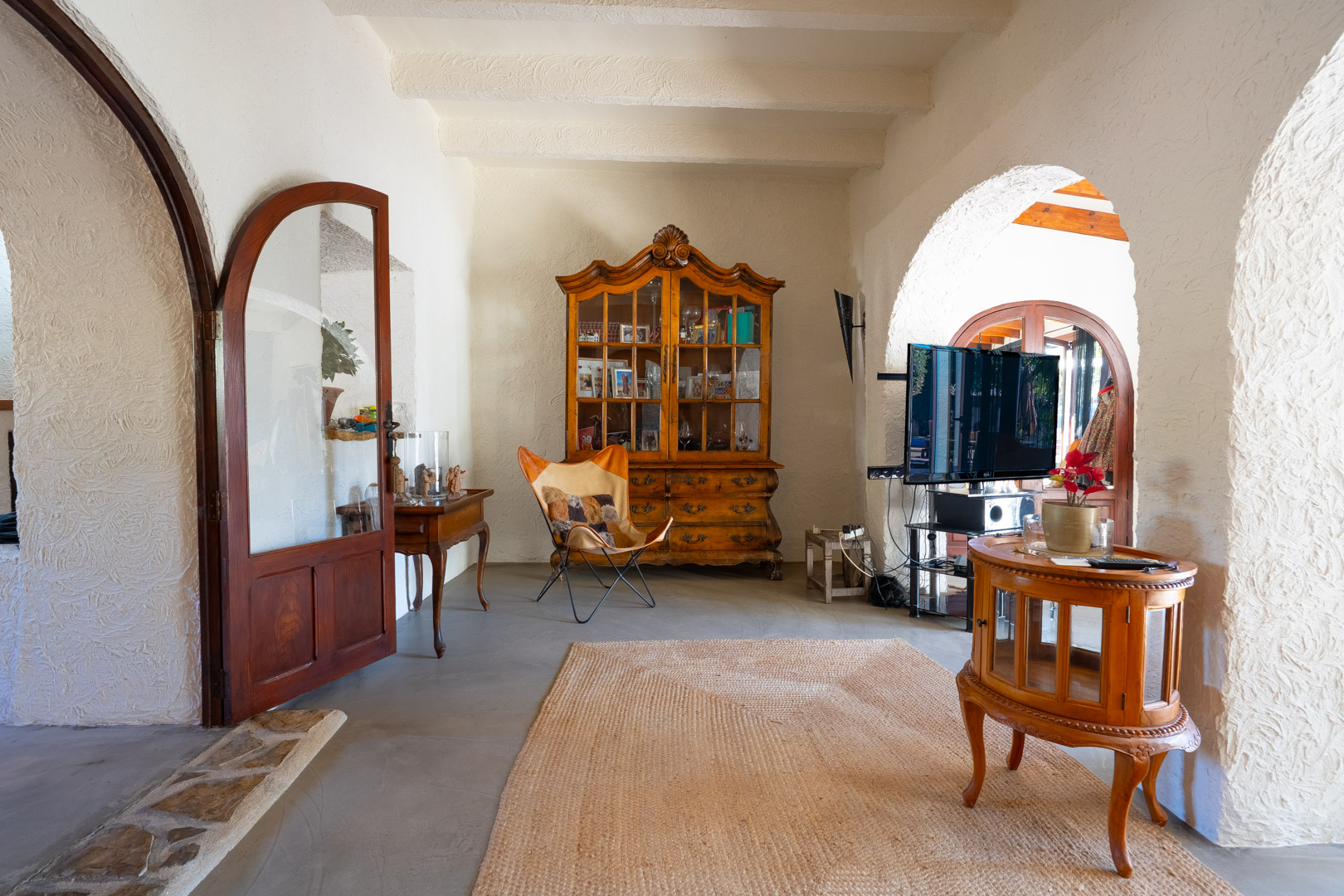 Sala de estar con techos de vigas blancas, arcos tradicionales y suelos de microcemento. Destaca una vitrina antigua de madera y mobiliario de estilo rústico.