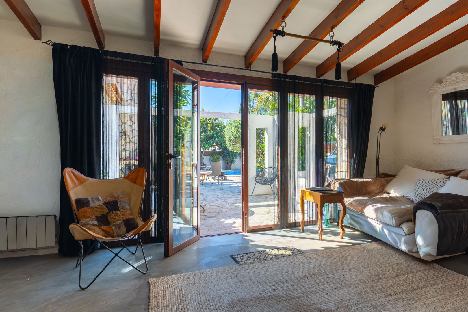 Salón luminoso con vigas de madera a la vista y grandes ventanales que conectan directamente con el patio empedrado y la zona de piscina.