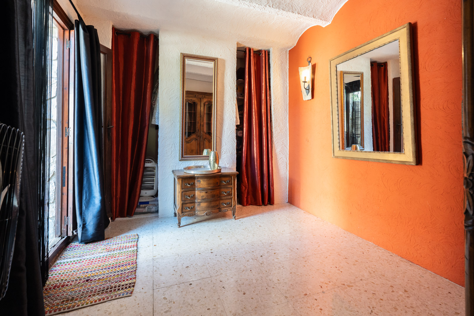 Zona de vestidor con pared de acento naranja, espejos ornamentados, cómoda de madera clásica y suelos de terrazo. Cortinas rojas resguardan los armarios empotrados.