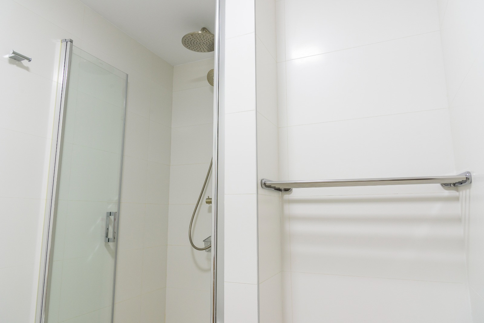 Baño moderno con zona de ducha, mampara de cristal y grifería cromada con rociador de efecto lluvia. Presenta revestimientos de azulejos blancos y un toallero metálico integrado.