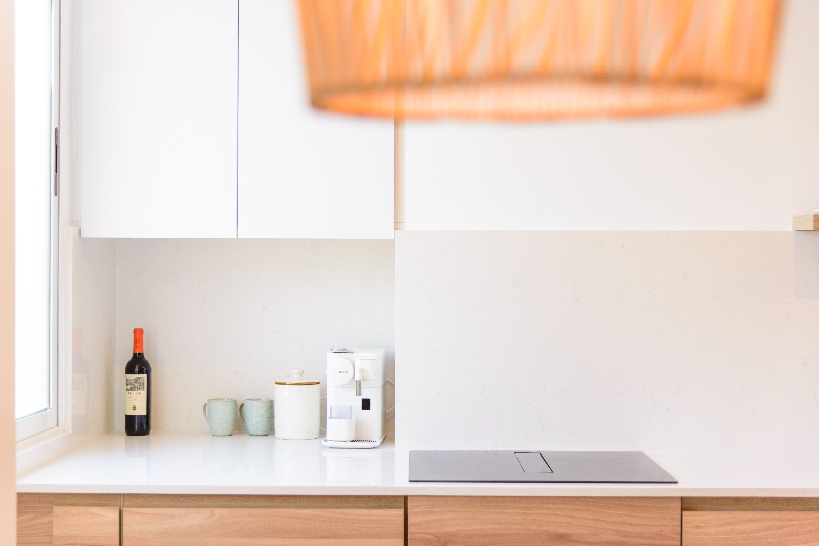Cocina de diseño con encimera blanca, placa de inducción integrada y armarios inferiores de madera. La luz natural lateral realza el salpicadero de piedra clara y el estilo minimalista.
