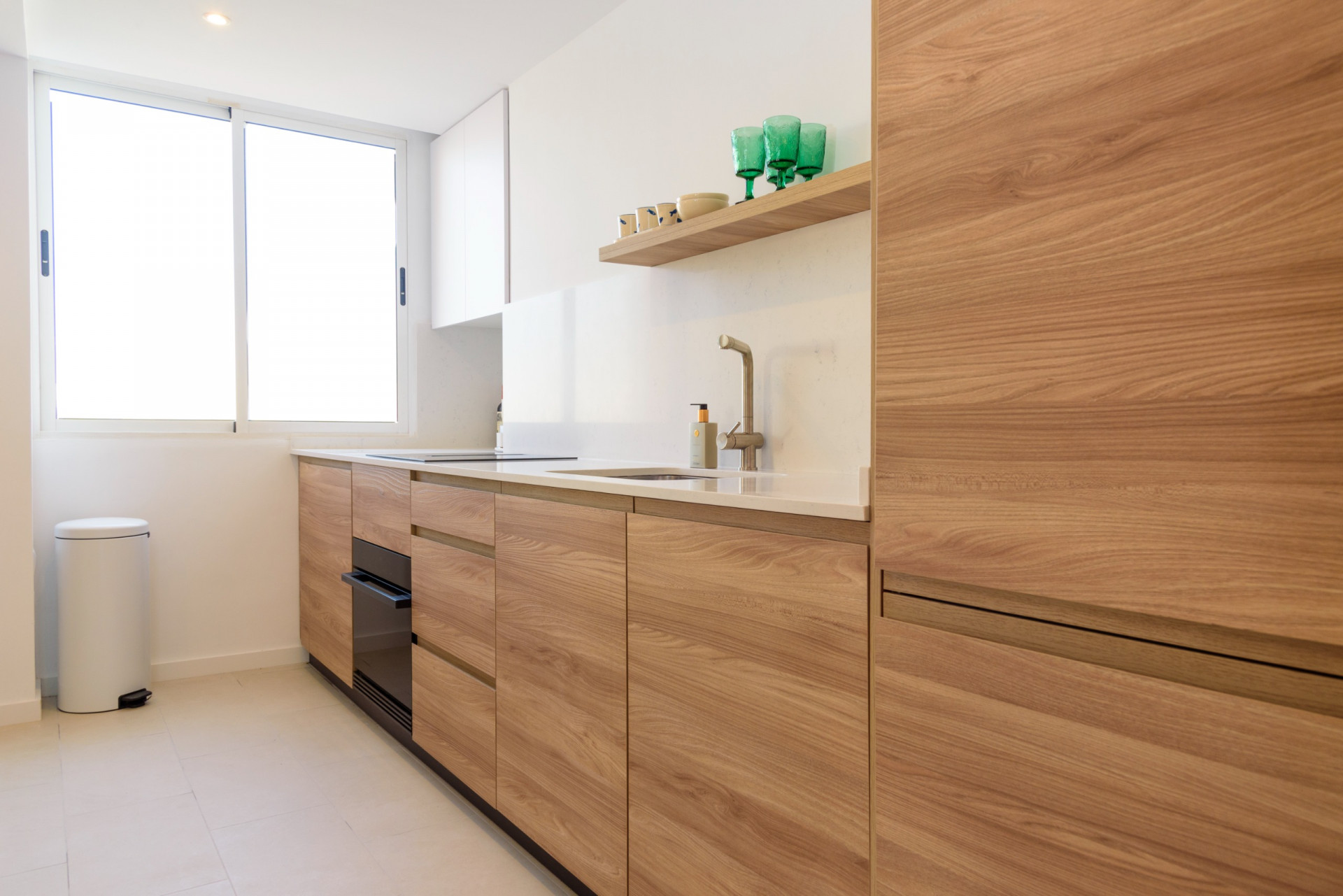 Cocina moderna con armarios de madera clara, encimera blanca y fregadero integrado. La luz natural entra por un gran ventanal, resaltando los acabados contemporáneos y funcionales.