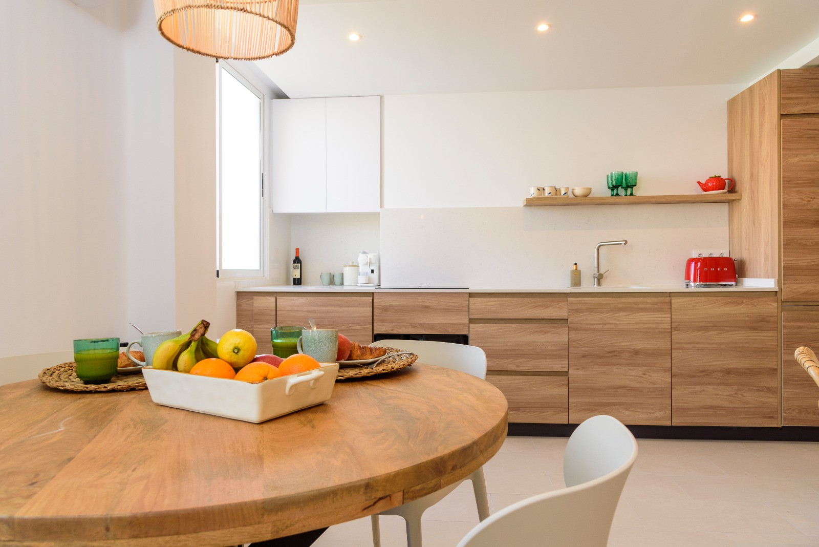 Comedor con mesa de madera circular y cocina integrada de diseño contemporáneo, equipada con armarios en acabado roble, encimera blanca y luz natural lateral.