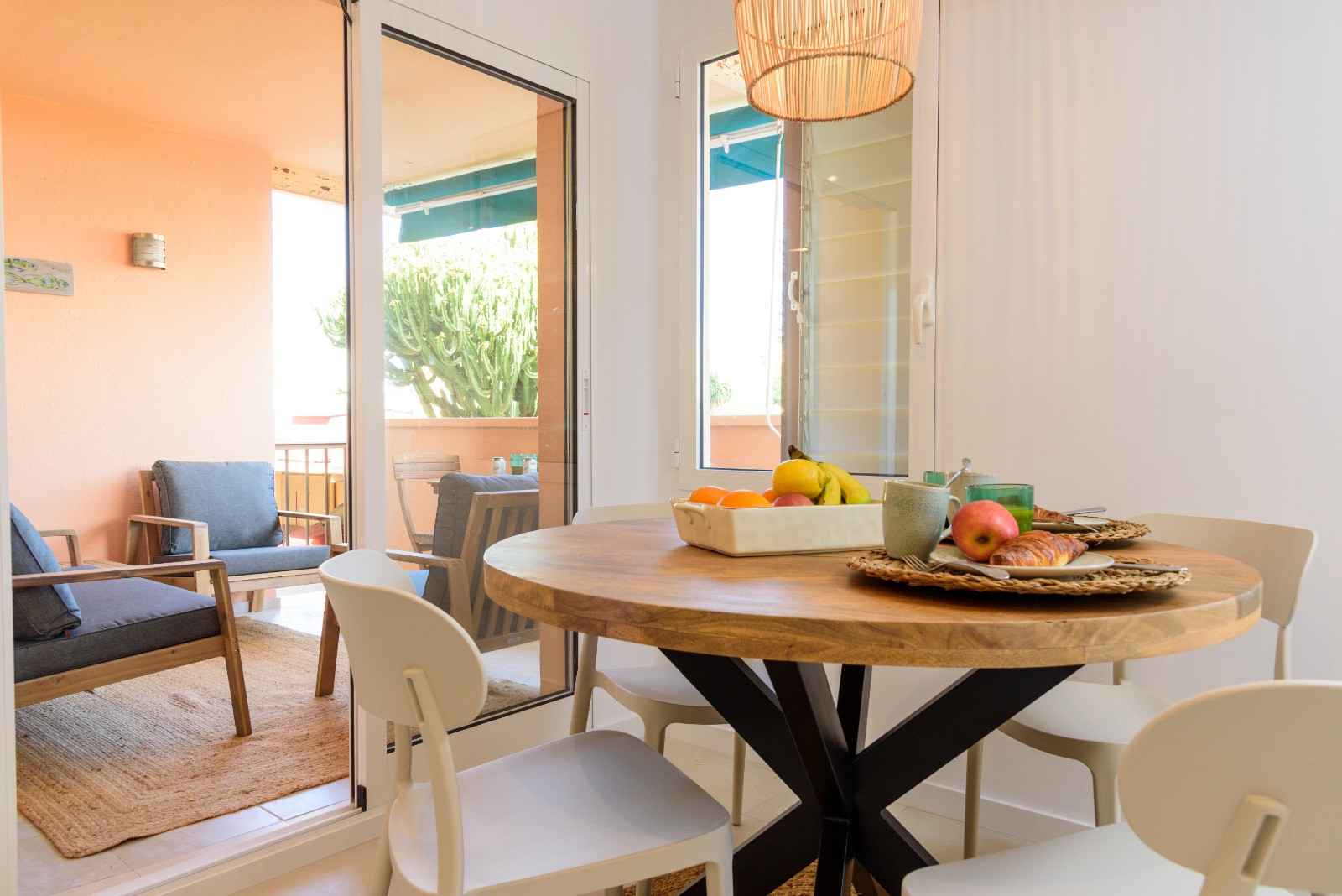 Comedor luminoso con mesa redonda de madera y sillas blancas, conectado a una terraza amueblada a través de grandes ventanales que maximizan la luz natural.