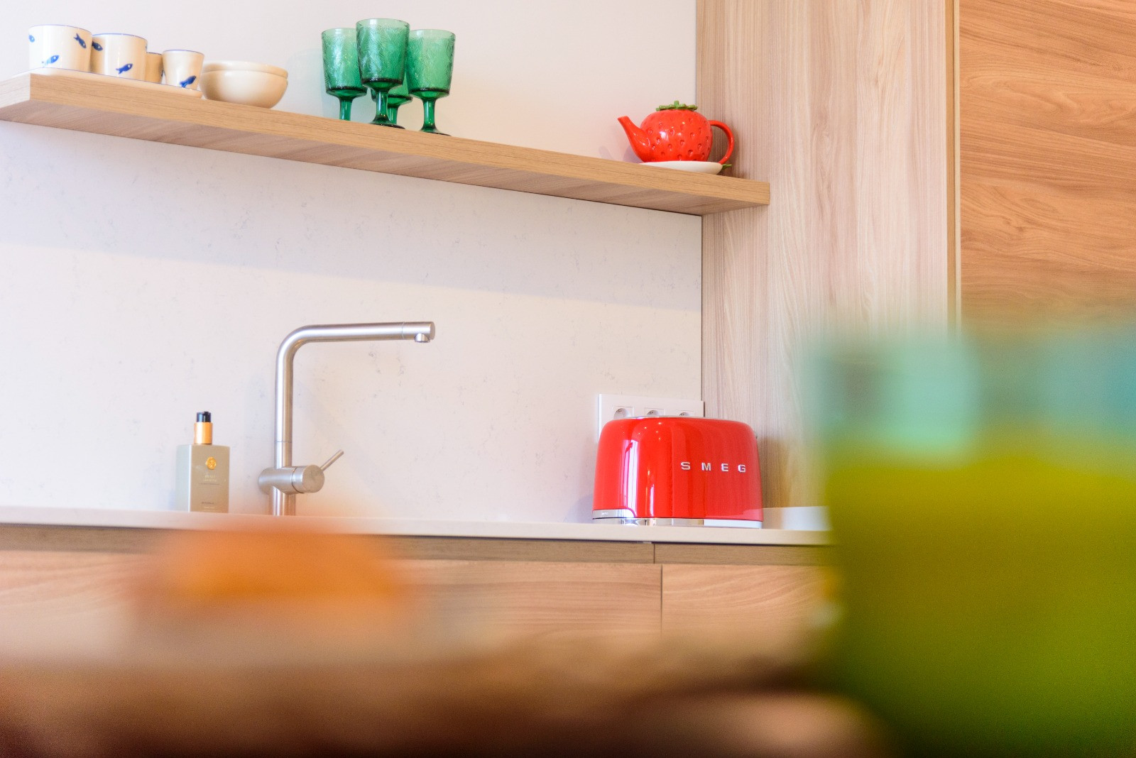 Detalle de la cocina con encimera blanca, grifería minimalista y estante de madera. Incluye electrodomésticos de diseño y acabados en tonos naturales.