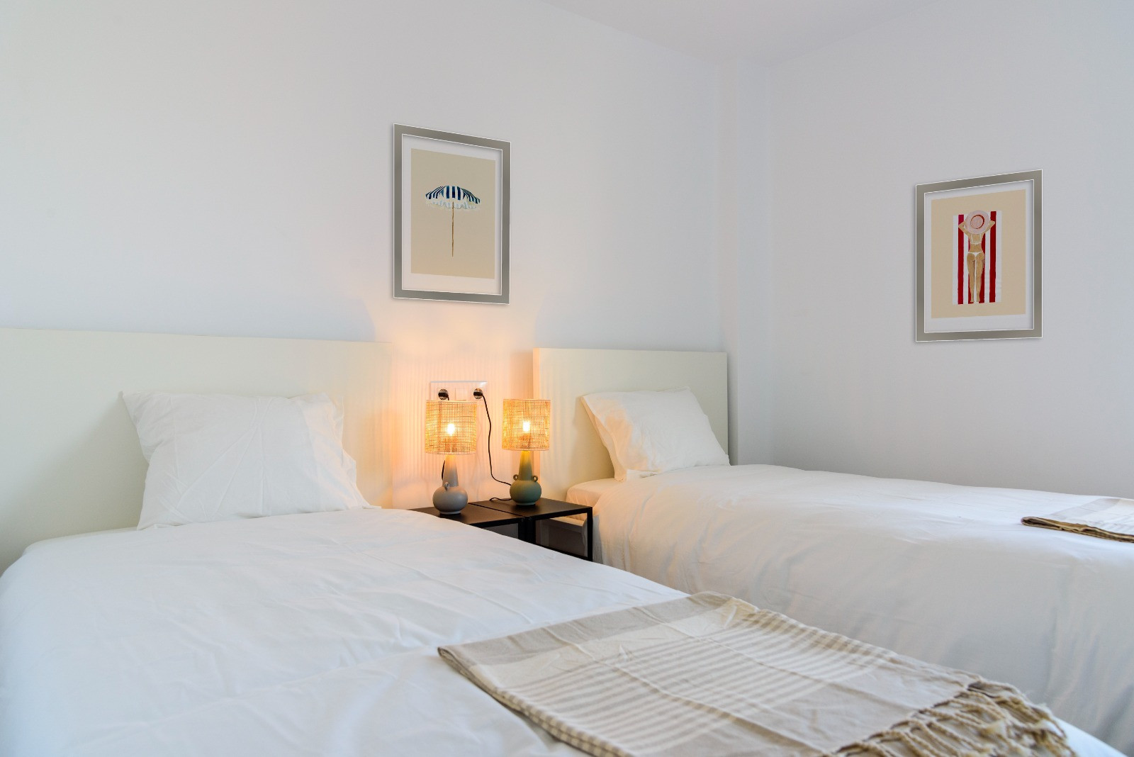 Dormitorio luminoso con dos camas individuales, cabeceros blancos y lámparas de fibra natural. Las paredes blancas están decoradas con cuadros de temática costera.